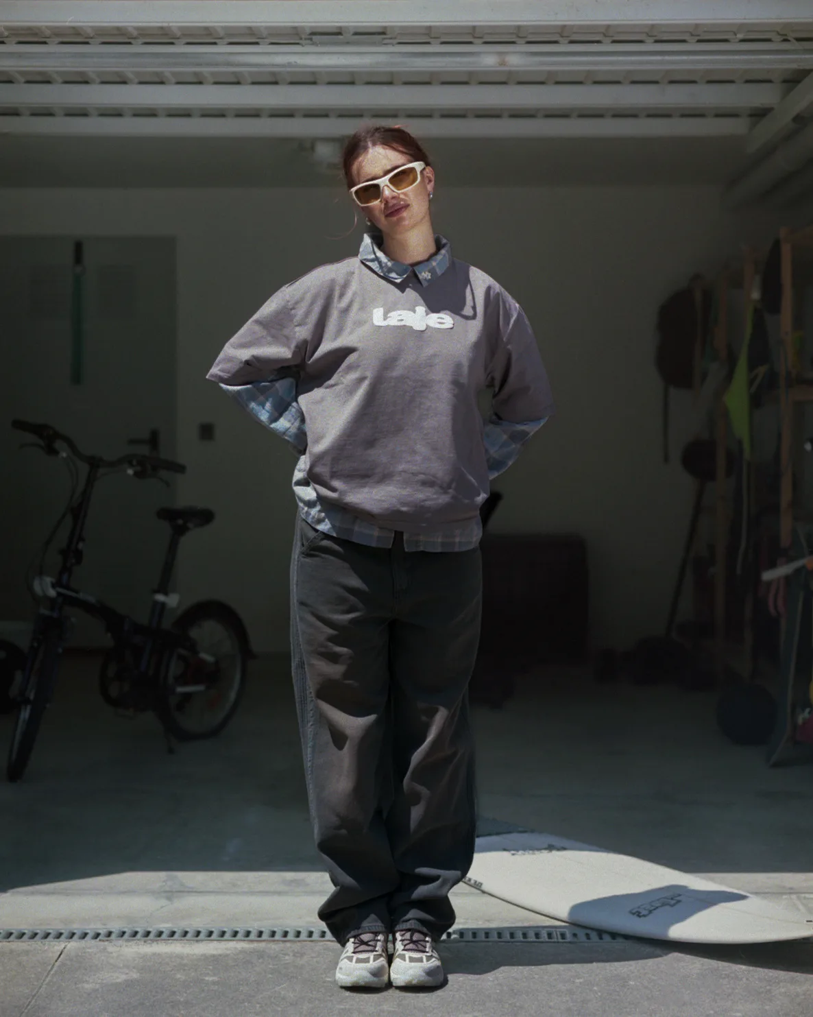 Woman in grey Laje sweatshirt and sunglasses, garage doorway, surfboard behind